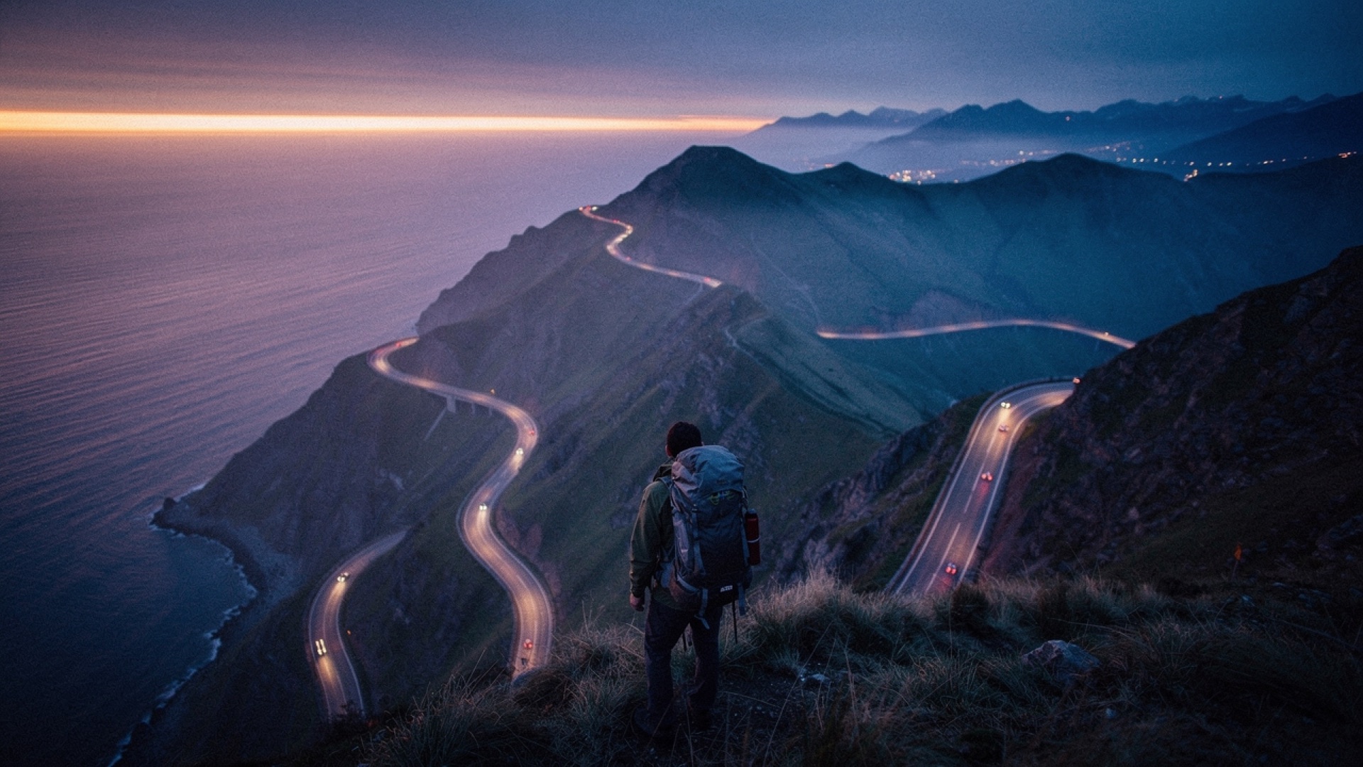 A backpacker on a ridge above a winding coastal road at dusk